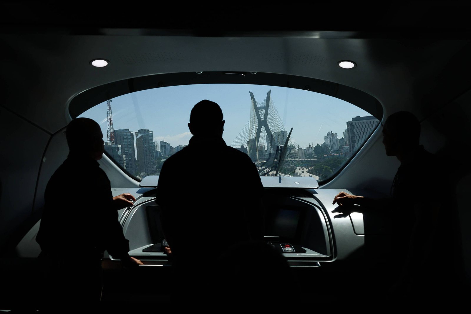 Três silhuetas de pessoas estão dentro de uma embarcação, olhando pela janela frontal para a Ponte Estaiada em São Paulo, com o horizonte urbano ao fundo em dia claro.