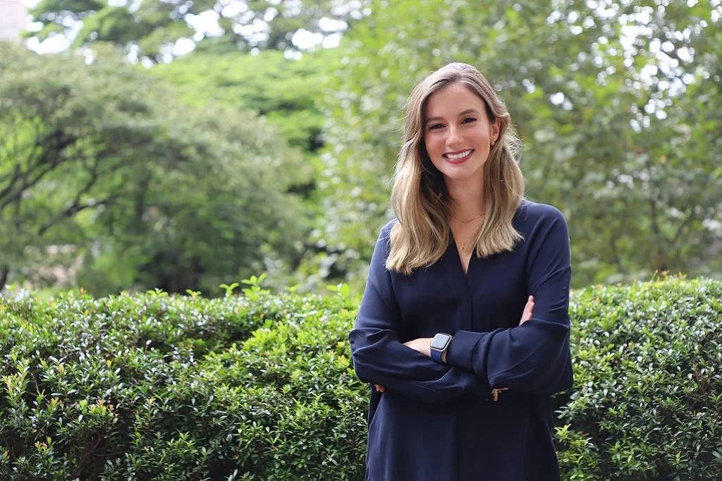 Mulher de cabelos loiros e lisos, vestindo blusa azul escura, está em pé com os braços cruzados diante de arbustos verdes. Ao fundo, árvores e vegetação formam um cenário natural.