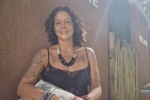 Mulher de cabelos cacheados e curtos sorri para a câmera, vestindo blusa preta de alças finas e segurando uma bolsa cinza. Ela está encostada em uma parede marrom com estrutura de bambu ao lado, sob luz natural.