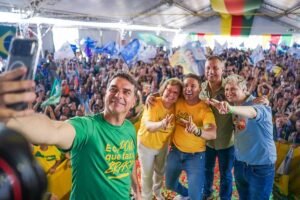 Quatro pessoas posam para selfie em primeiro plano durante evento político sob tenda. Multidão com bandeiras e cartazes ocupa o espaço ao fundo, com decoração nas cores verde, amarelo e vermelho no teto.