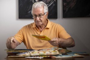 Homem idoso de cabelos brancos e óculos, vestindo camisa polo laranja, sentado à mesa organizando livros infantis coloridos espalhados à sua frente.