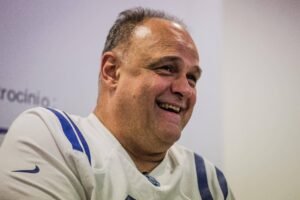 Homem de meia-idade com cabelo curto e ralo sorri olhando para a direita. Ele veste uma camisa esportiva branca com detalhes azuis e um logo da Nike na manga esquerda. O fundo é neutro e desfocado.