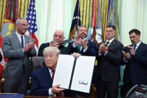 Ex-presidente dos Estados Unidos sentado na mesa do Salão Oval, segurando documento assinado. Cinco homens em pé ao fundo aplaudem, com bandeiras americanas e militares atrás.