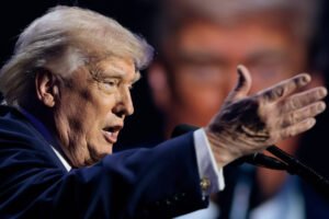 Ex-presidente dos Estados Unidos fala ao microfone com gesto de mão estendida à frente. Fundo desfocado mostra imagem ampliada do rosto dele.