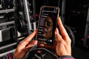 Mãos com munhequeiras seguram celular exibindo texto de plano de treino para a próxima semana. Fundo mostra equipamentos de musculação em ambiente de academia.