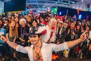 Cosplayer agachado no centro do palco com camisa branca aberta e chapéu preto com pena, fazendo gesto com os braços. Ao fundo, grande público sorridente em ambiente interno iluminado, com estandes e painéis coloridos.
