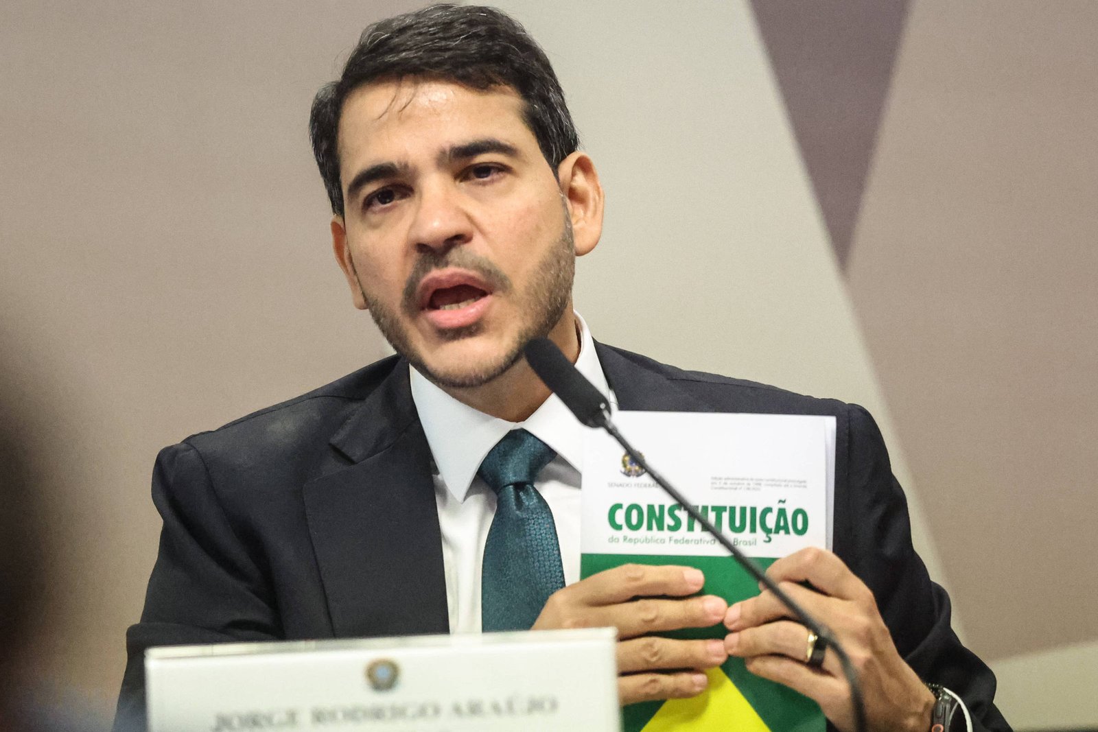Homem de terno escuro e gravata verde fala ao microfone enquanto segura um exemplar da Constituição do Brasil. Placa com nome e cargo está à sua frente em mesa de audiência.