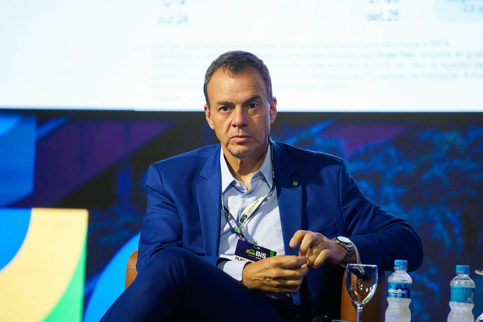 Homem de terno azul sentado em cadeira, segurando microfone, com copo e garrafas de água sobre mesa ao lado. Fundo apresenta cores azul, amarelo e branco.