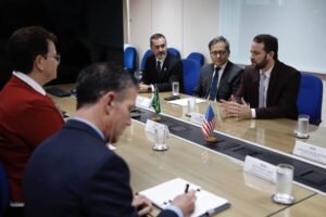 Cinco homens sentados ao redor de uma mesa de conferência discutem assuntos formais. Dois deles estão de um lado da mesa, um deles fala gesticulando, enquanto os outros três escutam atentamente. Pequenas bandeiras do Brasil e dos Estados Unidos estão posicionadas no centro da mesa. Copos de água e cadernos estão sobre a mesa, e cadeiras azuis compõem o ambiente.