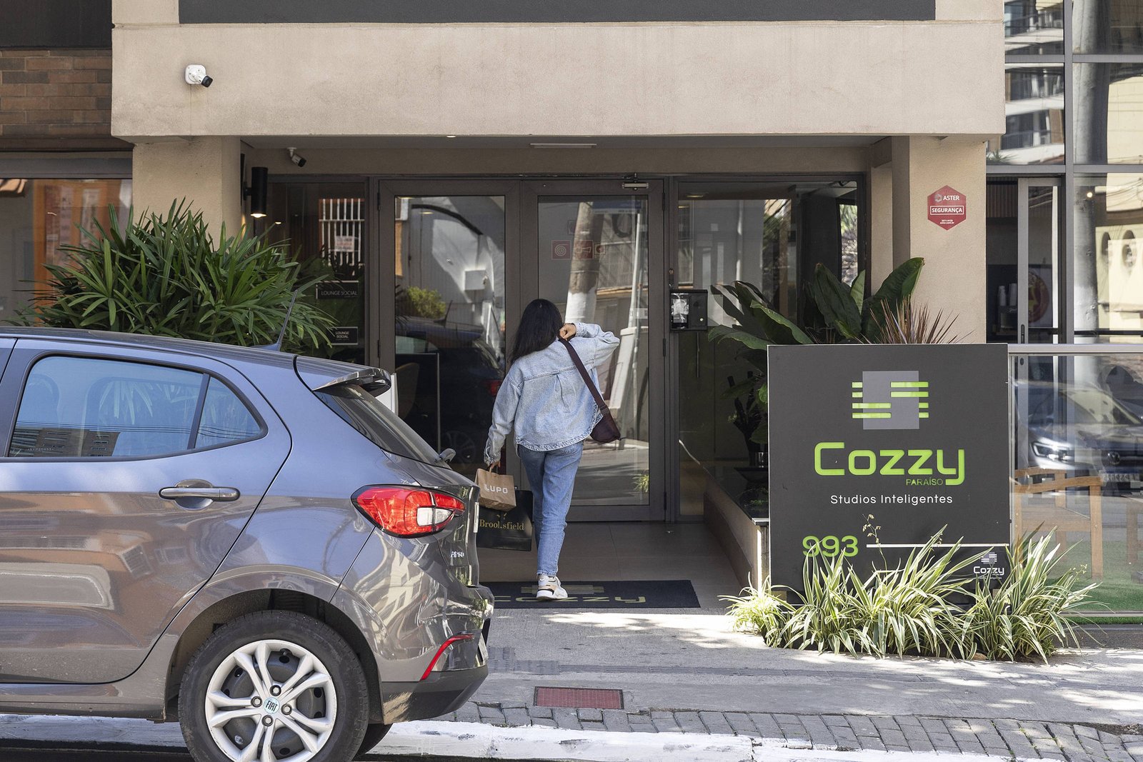 Pessoa de costas, vestindo jaqueta jeans e calça azul, entra na porta de vidro de um prédio comercial com placa