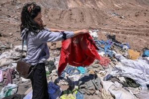 Mulher de cabelos escuros e roupas casuais segura uma peça vermelha enquanto observa um grande amontoado de roupas usadas espalhadas sobre solo seco e rochoso.