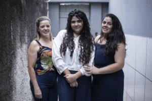 Três mulheres estão lado a lado em um corredor estreito com paredes cinza. A mulher à esquerda usa blusa estampada e calça jeans, a do meio veste camisa branca e calça escura, e a da direita usa vestido azul escuro. Todas sorriem para a câmera.