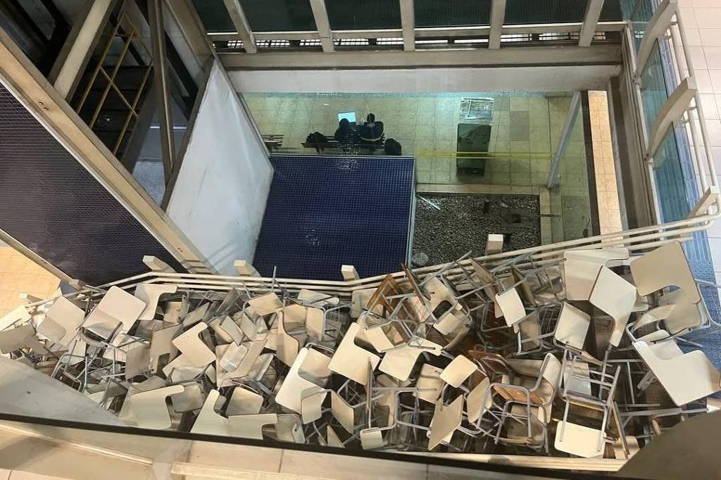 Grande quantidade de cadeiras escolares empilhadas desordenadamente em área interna de prédio, vista de cima. Ao fundo, duas pessoas sentadas em bancos próximos a uma parede de vidro.