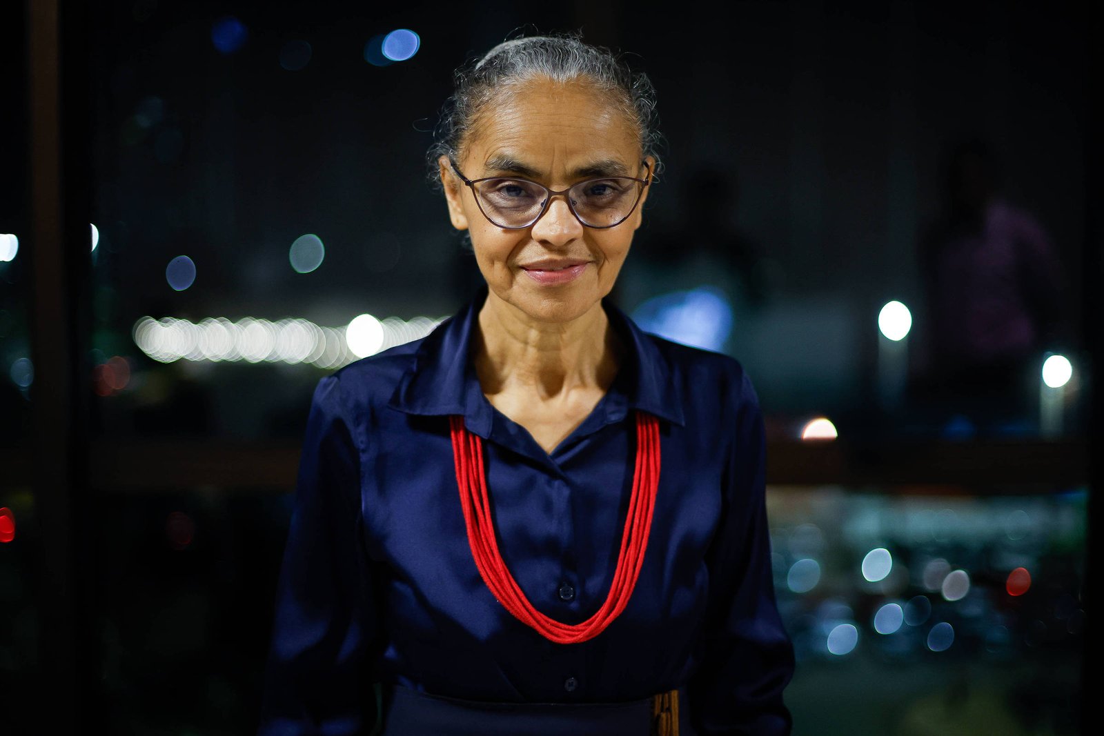 Mulher idosa com cabelo grisalho preso, usando óculos e camisa azul escura, veste colar vermelho em ambiente com luzes desfocadas ao fundo à noite.