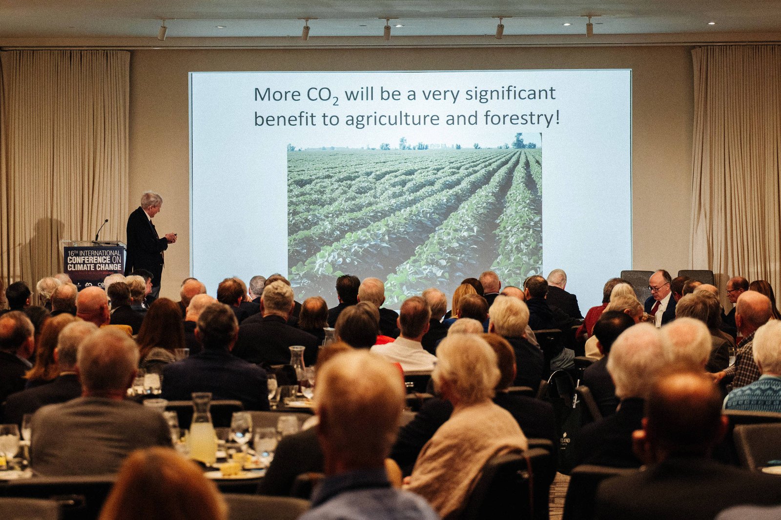 Homem apresenta palestra para público sentado em auditório. Tela exibe texto