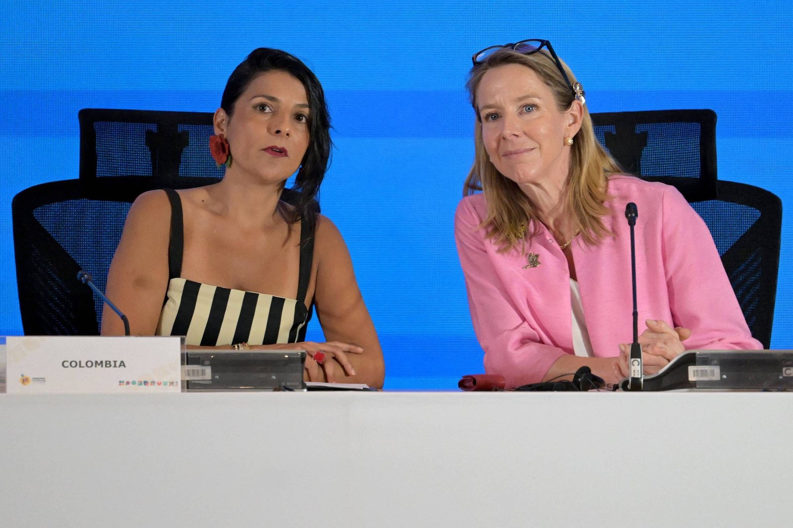 Duas mulheres sentadas lado a lado em uma mesa branca com microfones. A mulher à esquerda usa vestido listrado preto e branco e brincos vermelhos, com placa indicando