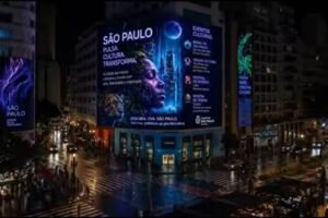 Painel digital iluminado em fachada curva de edifício no centro de São Paulo exibe campanha cultural com rosto feminino e texto sobre eventos culturais. Ruas molhadas refletem luzes da cidade e trânsito de veículos e pedestres é visível à noite.