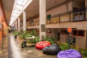 Corredor interno amplo com piso de pedra, plantas ao longo do centro e três pufes coloridos (vermelho, preto e roxo) próximos a uma mesa pequena. À esquerda, paredes com portas e janelas de vidro, e ao fundo uma pessoa caminhando. No lado direito, mezanino com corrimão azul e placas informativas nas paredes.