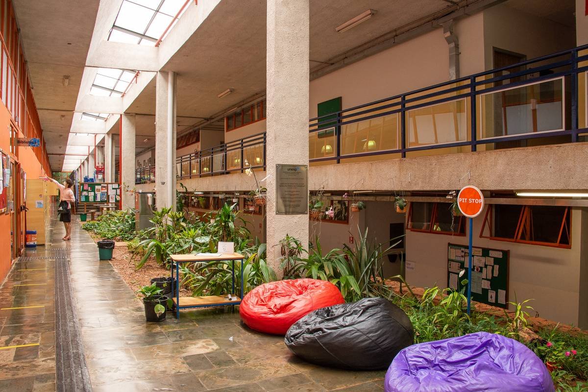 Corredor interno amplo com piso de pedra, plantas ao longo do centro e três pufes coloridos (vermelho, preto e roxo) próximos a uma mesa pequena. À esquerda, paredes com portas e janelas de vidro, e ao fundo uma pessoa caminhando. No lado direito, mezanino com corrimão azul e placas informativas nas paredes.