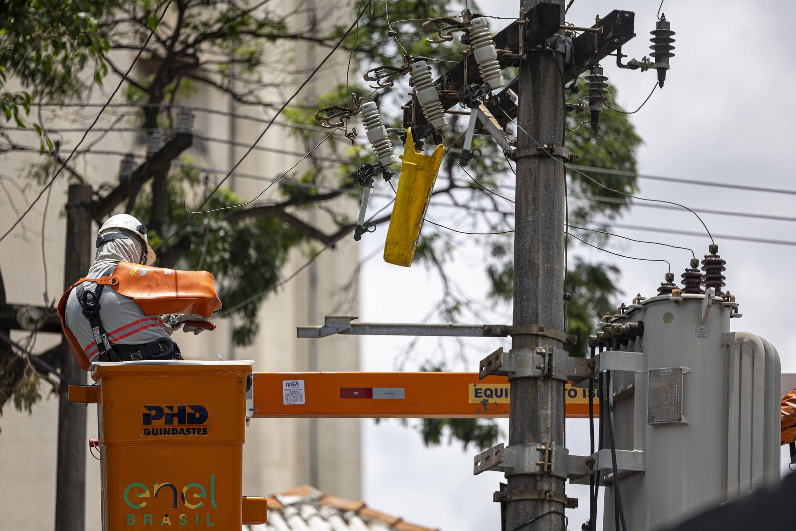 Técnico com capacete e colete laranja opera em plataforma elevatória próxima a transformador em poste de energia. Fios e isoladores visíveis, ambiente urbano com árvores e edifícios ao fundo.