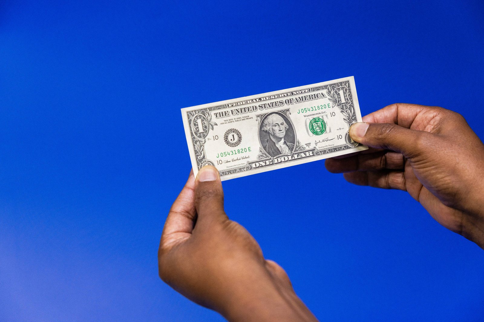 A imagem mostra uma nota de um dólar sendo segurada por duas mãos. O fundo é azul. A nota apresenta a figura de George Washington e o texto