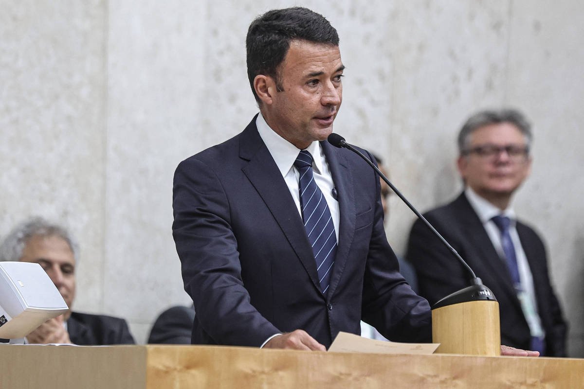 Um homem em um terno escuro está falando em um microfone em um evento oficial. Ele está posicionado atrás de um púlpito de madeira e parece estar fazendo um discurso. Ao fundo, há outras pessoas sentadas, algumas delas olhando para ele. A parede atrás é clara e simples, e há um dispositivo eletrônico à esquerda do púlpito.