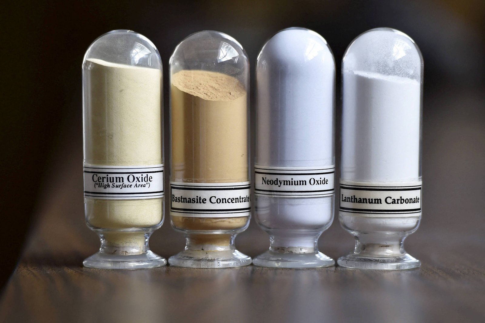 Quatro frascos transparentes alinhados contendo pós de diferentes cores e texturas. Da esquerda para a direita: pó amarelo claro rotulado como Cerium Oxide, pó marrom claro rotulado como Bastnasite Concentrate, pó branco rotulado como Neodymium Oxide e pó branco rotulado como Lanthanum Carbonate. Fundo desfocado e superfície de madeira escura.