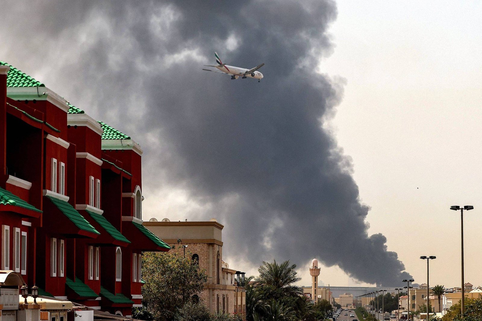 Avião comercial voa sobre área urbana com prédios vermelhos e verdes à esquerda. Ao fundo, grande nuvem de fumaça preta se eleva no céu claro, indicando possível incêndio ou explosão. Rua com postes de iluminação e vegetação visível na cena.