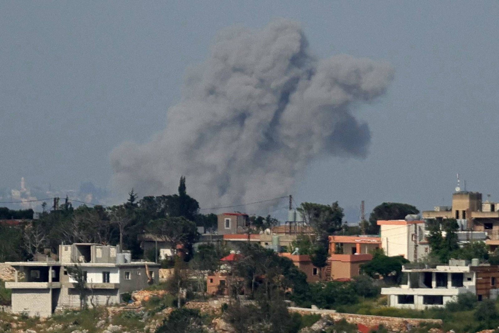 Nuvem espessa de fumaça cinza escuro se eleva atrás de casas em área urbana com vegetação dispersa. Céu claro ao fundo.