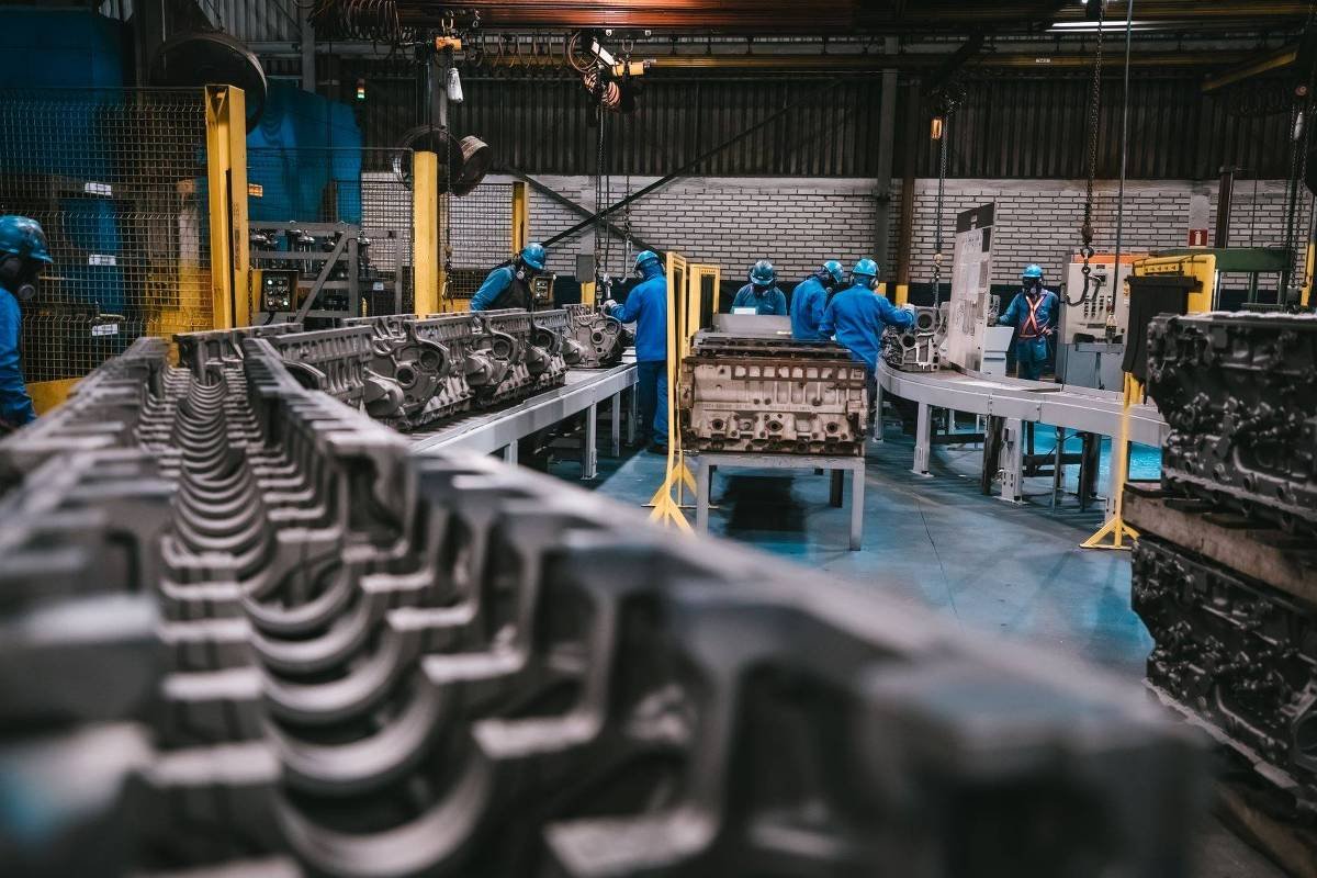 Funcionários com uniformes azuis e capacetes trabalham em linha de montagem dentro de fábrica industrial. Grandes moldes metálicos estão dispostos em bancadas ao redor do ambiente.