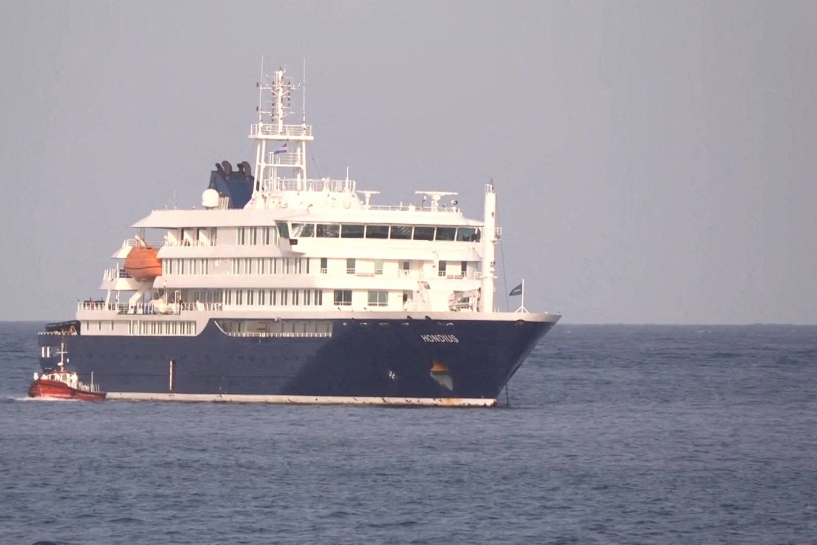 Navio de passageiros branco e azul escuro navegando em mar calmo sob céu claro. Barco menor vermelho acompanha ao lado esquerdo do navio maior.