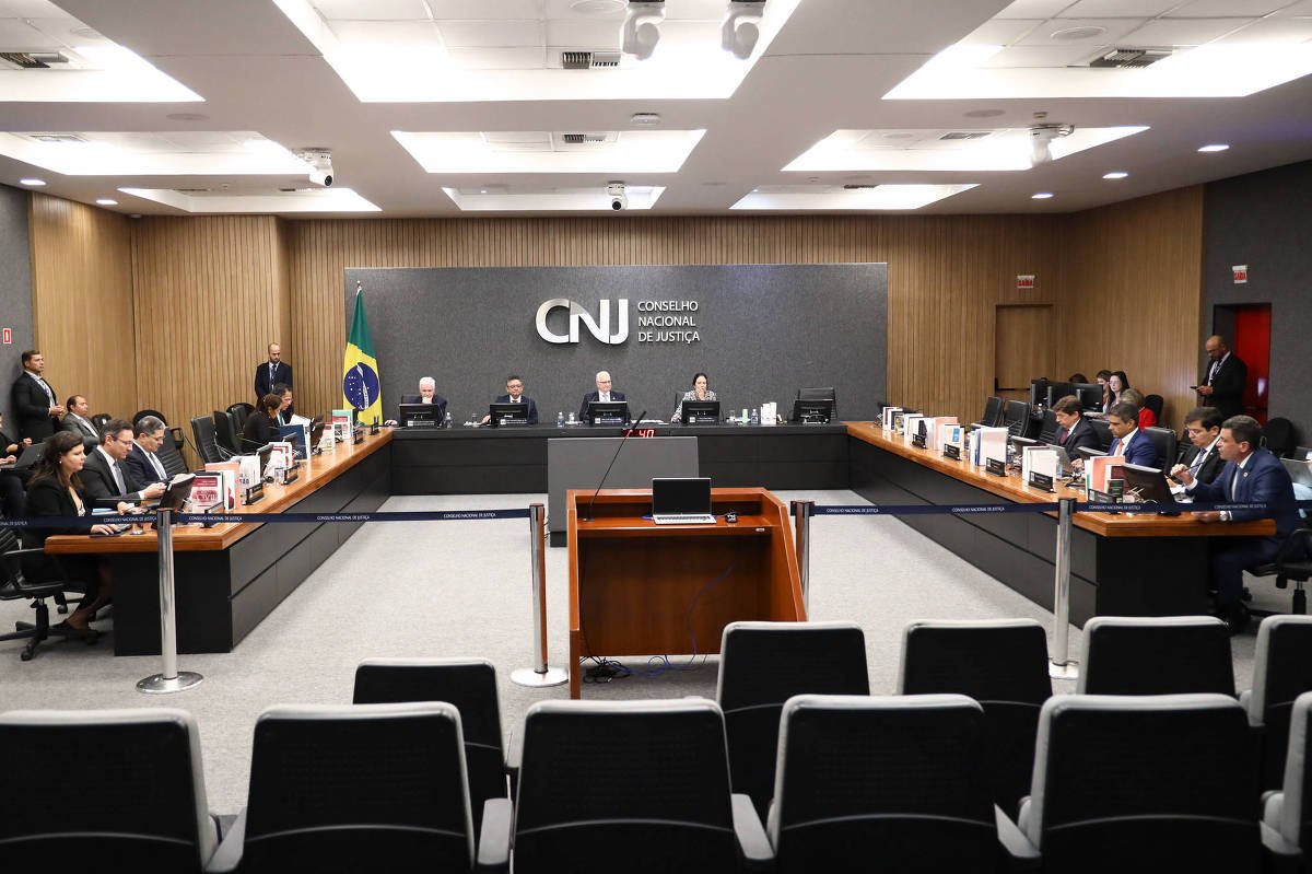 Plenário do Conselho Nacional de Justiça com membros sentados em mesas em formato de U. Bandeira do Brasil ao fundo e painel com logo do CNJ. Ambiente formal com cadeiras vazias em primeiro plano.