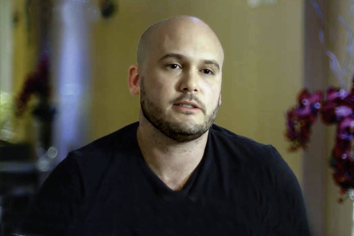 Homem careca com barba curta e camiseta preta sentado em ambiente interno com fundo desfocado que inclui flores vermelhas.