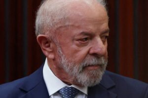 Homem idoso com cabelo branco e barba grisalha veste terno azul escuro, camisa branca e gravata azul com padrão. Fundo com painéis verticais escuros.