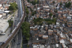 Vista aérea mostra favela com casas pequenas e amontoadas ao lado de linha de trem elevada. Trem branco circula na via férrea paralela a uma avenida com poucos carros. Vegetação dispersa aparece entre as construções e ao longo da linha férrea.