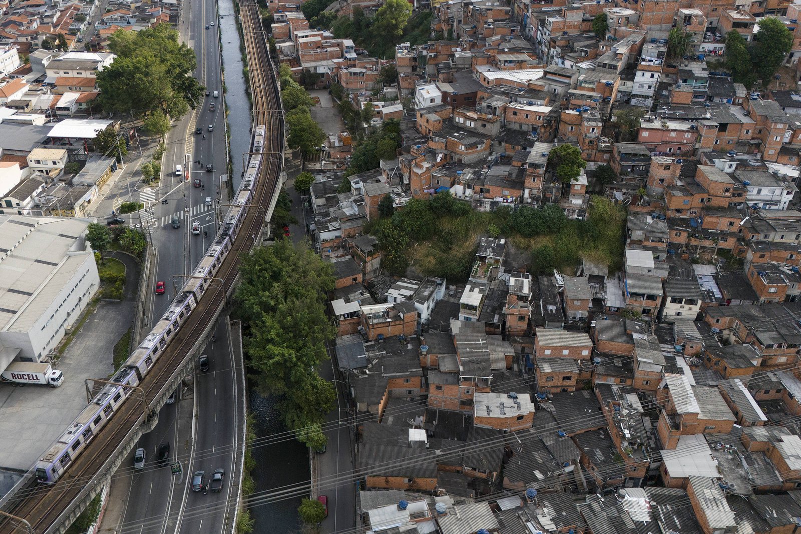 Vista aérea mostra favela com casas pequenas e amontoadas ao lado de linha de trem elevada. Trem branco circula na via férrea paralela a uma avenida com poucos carros. Vegetação dispersa aparece entre as construções e ao longo da linha férrea.