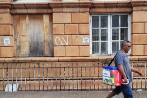 Um homem caminha pela calçada, carregando uma bolsa térmica colorida. Ao fundo, há uma parede de um edifício com uma janela de vidro e outra com madeira coberta no lugar da janela, além de grafites visíveis na parede. Um deles tem inscritas as letras CV. O homem está vestido com uma camiseta cinza e calças escuras.