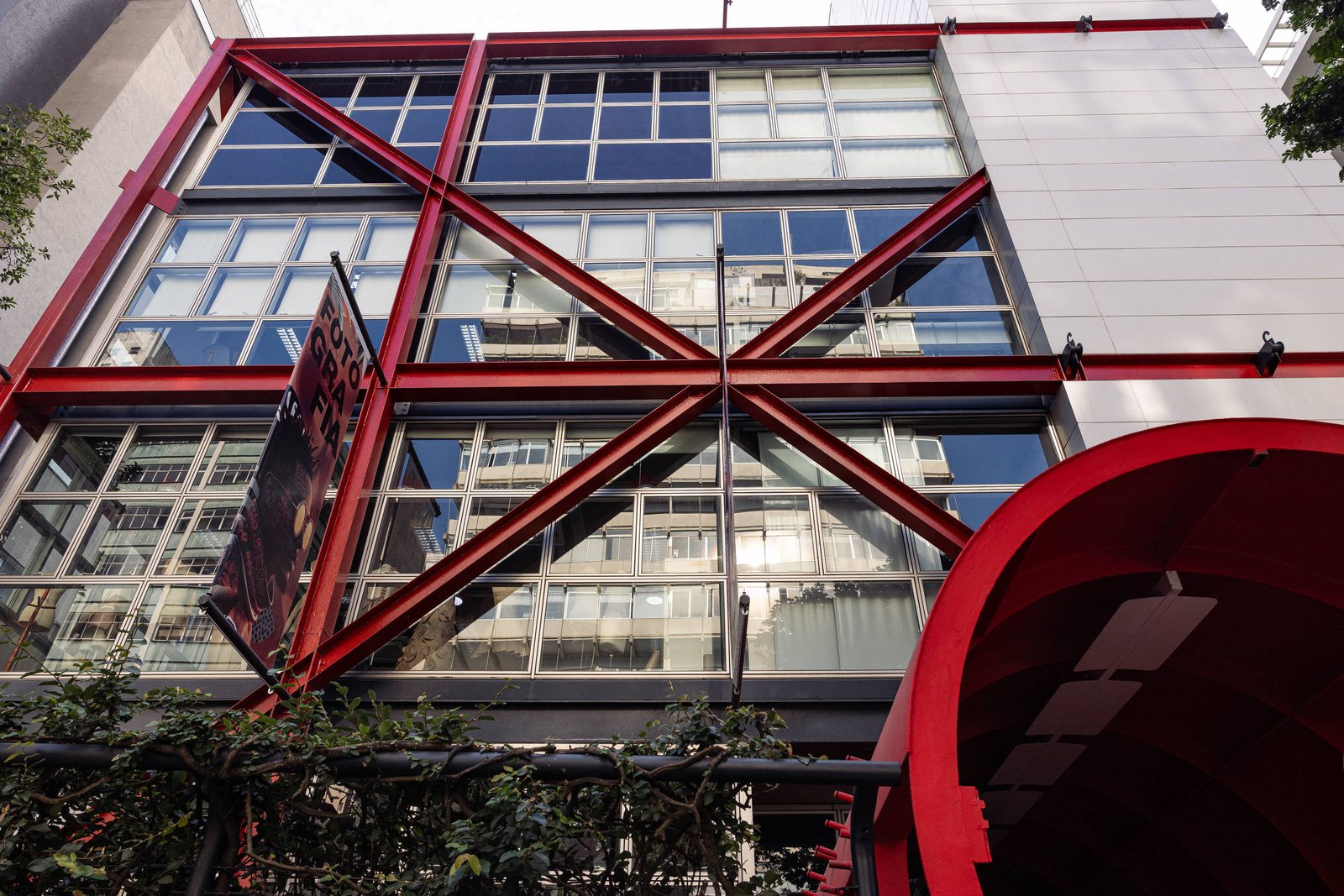 Prédio com fachada de janelas amplas refletindo edifícios vizinhos, sustentada por estrutura metálica vermelha em formato de X. Entrada com arco vermelho à direita e vegetação na base.
