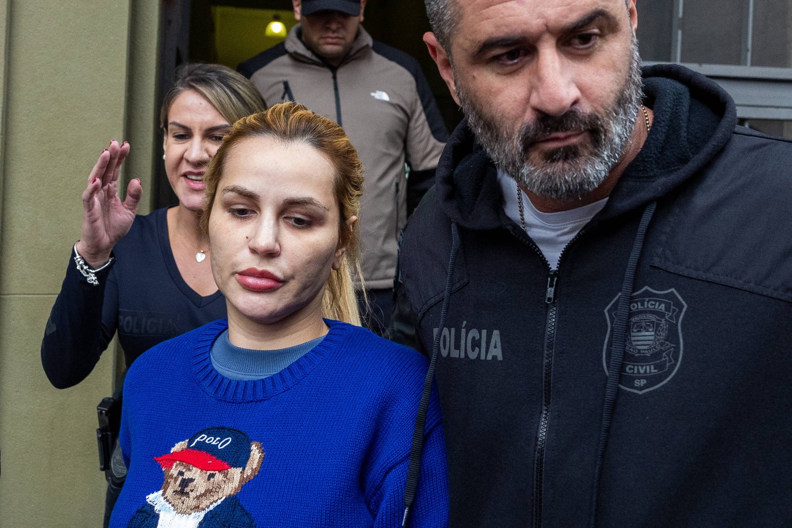Mulher de cabelo loiro preso e suéter azul com estampa de urso é escoltada por dois policiais civis, um homem à frente e uma mulher atrás, ao sair de um prédio.