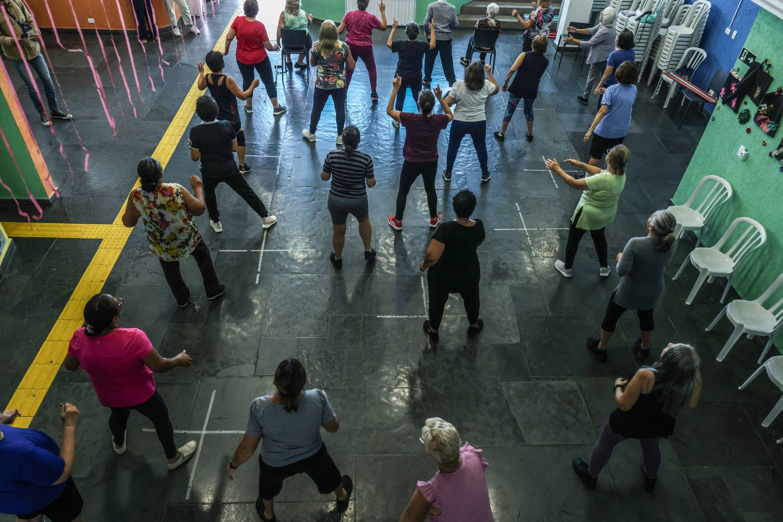 Vinte e uma pessoas, majoritariamente mulheres, estão distribuídas em um salão com piso escuro, realizando exercícios físicos em posições semelhantes, com braços levantados. O ambiente tem cadeiras brancas empilhadas e paredes coloridas, com faixas amarelas no chão delimitando espaços.