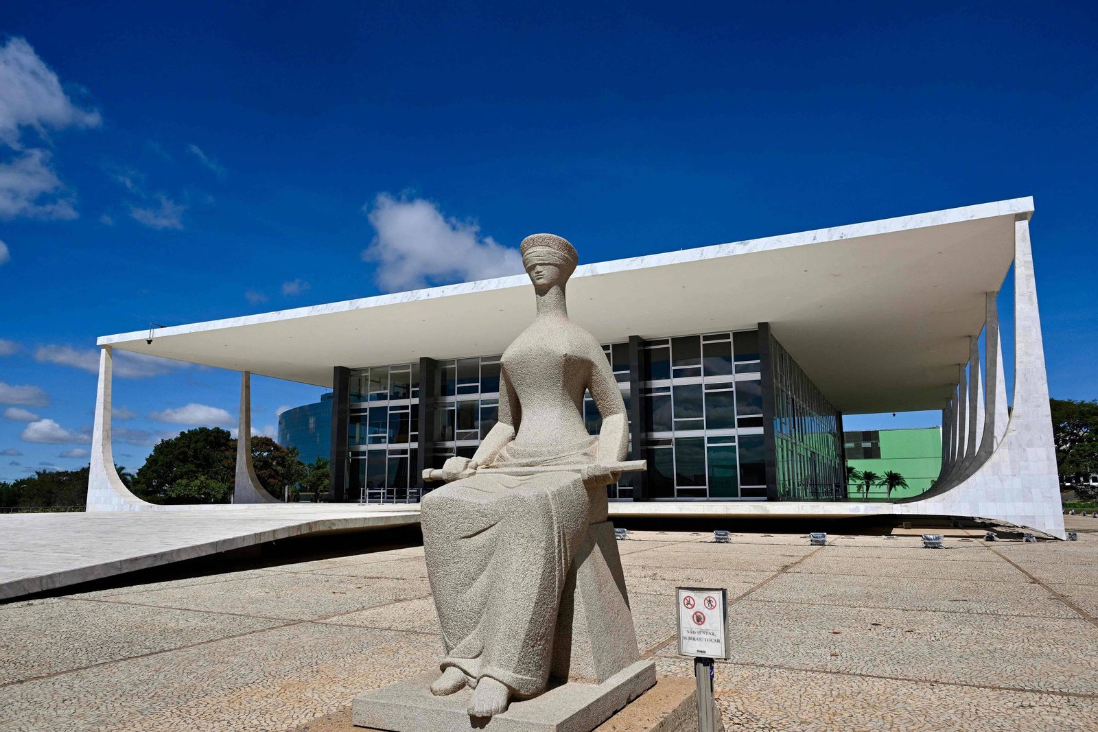 A imagem mostra o Supremo Tribunal Federal (STF) do Brasil, um edifício moderno com uma estrutura de concreto e vidro. Em primeiro plano, há uma escultura de uma figura feminina, representando a Justiça, que está sentada e usa uma venda nos olhos. O céu está claro e azul, com algumas nuvens.