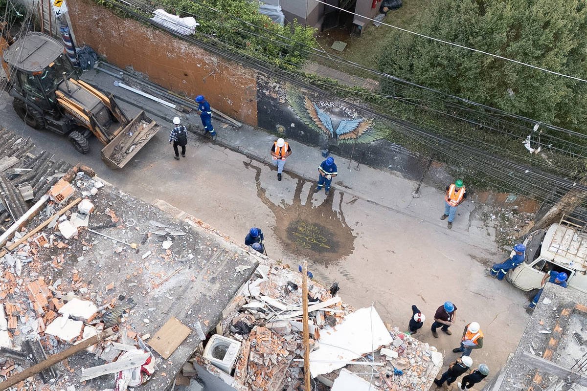 Vista aérea mostra destroços de um edifício demolido com escombros espalhados. Operários com coletes refletores e capacetes estão no entorno, junto a uma retroescavadeira estacionada na rua ao lado.