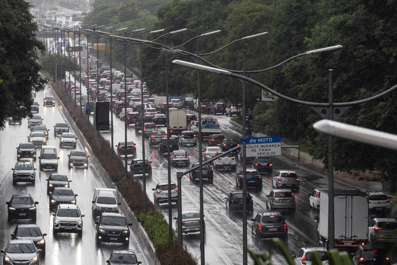 Congestionamento intenso na Marginal Pinheiros com veículos parados e lentos em várias faixas. Chuva deixa a pista molhada e reflexos visíveis no asfalto. Árvores e postes de iluminação acompanham a via.