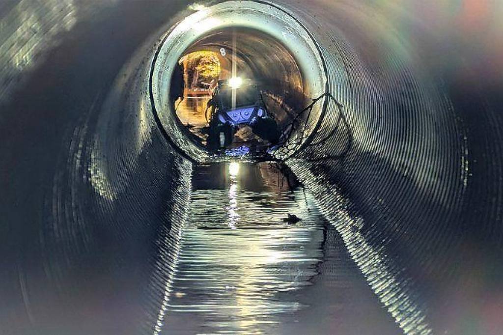 Pessoa usando capacete e equipamento de proteção está dentro de um tubo cilíndrico grande e metálico, parcialmente cheio de água. A luz ilumina o interior do tubo, destacando a superfície ondulada da água e as paredes internas do tubo.