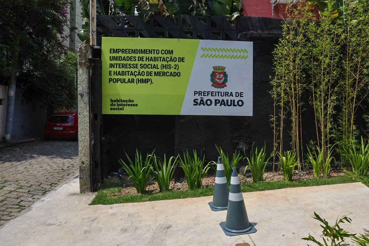 Placa verde e branca da Prefeitura de São Paulo anuncia empreendimento com unidades de habitação de interesse social e mercado popular, fixada em muro preto. Em frente, cones de sinalização e plantas ornamentais em área pavimentada e jardim com bambus ao lado.