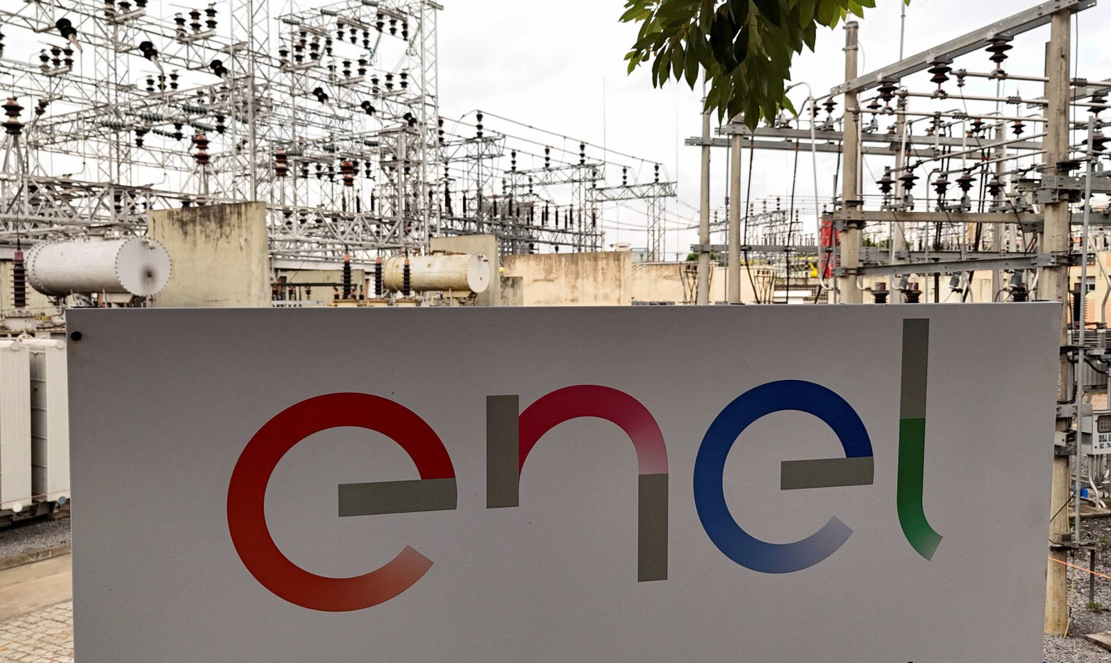 A imagem mostra uma placa da empresa Enel em primeiro plano, com o logotipo colorido da empresa. Ao fundo, há uma instalação elétrica com várias estruturas metálicas e equipamentos, como transformadores e cabos, sob um céu nublado.