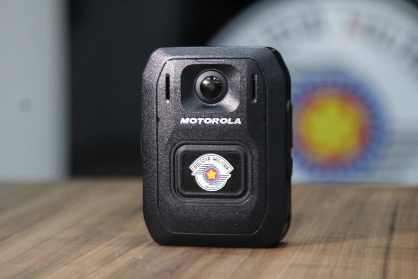 A imagem mostra uma câmera de polícia da marca Motorola, com um design robusto e uma lente na parte frontal. A câmera possui um emblema ou adesivo na parte inferior, que pode indicar a afiliação a uma força policial. O fundo é desfocado, mas parece haver um logotipo ou símbolo relacionado à polícia.