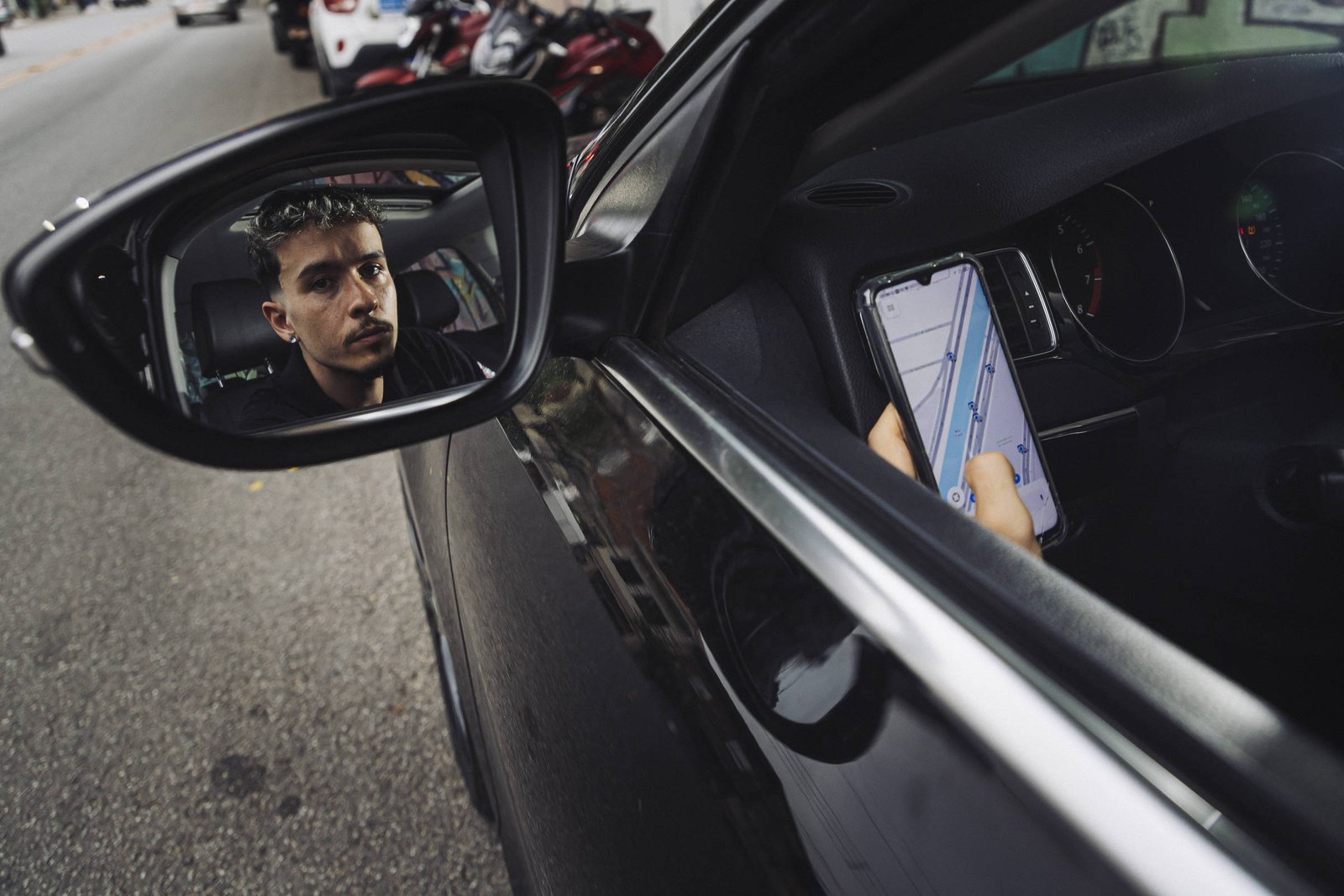 Homem jovem aparece refletido no retrovisor lateral de carro preto estacionado na rua. Ele segura um celular com aplicativo de navegação aberto, visível pela janela do motorista.