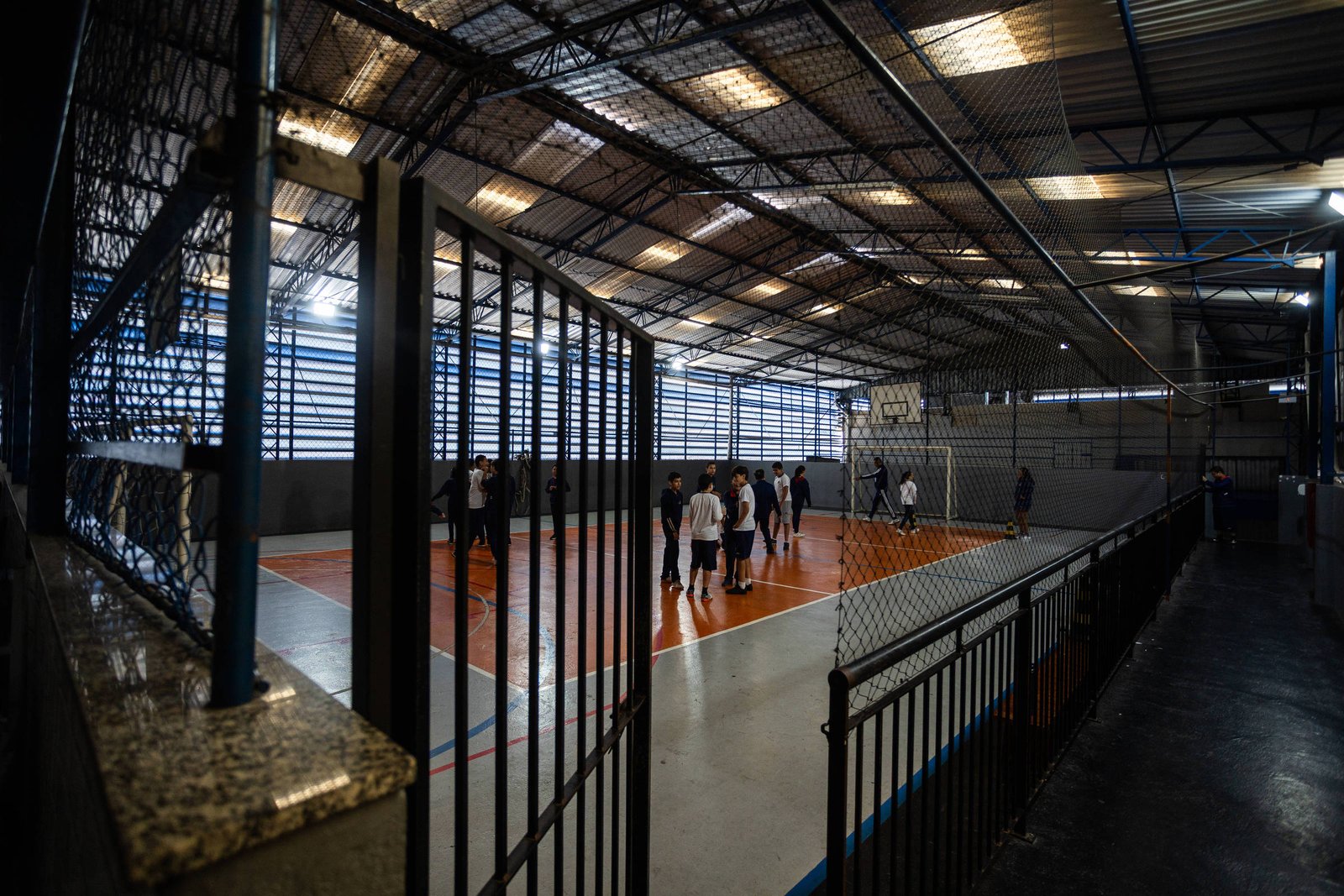 Grupo de pessoas jogando basquete em quadra coberta com piso laranja e branco. Estrutura metálica e iluminação natural por janelas laterais.