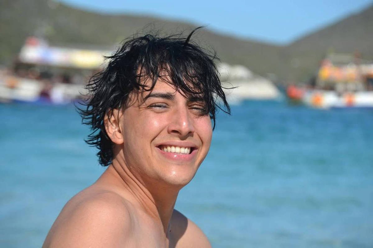 Jovem de cabelos escuros e molhados sorri para a câmera na praia. Ao fundo, mar azul e barcos coloridos próximos à costa com colinas ao longe.